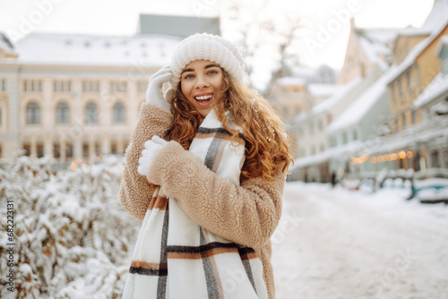 Young happy woman in a winter coat and scarf walks along a snowy city street. Street photo of a fashionable tourist enjoying the weather outdoors. Concept of style, weekend.