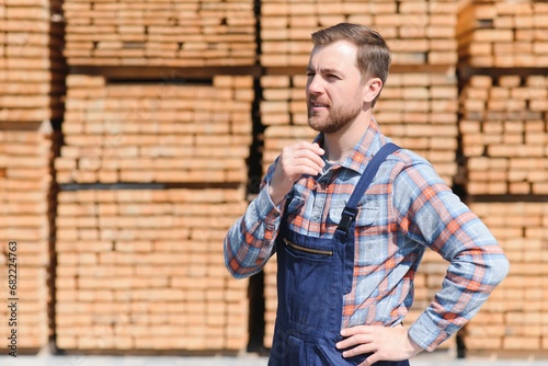 Portrait of a handsome worker choosing the best wooden boards. Carpenter standing next to a big stack of wood bars in a warehouse.