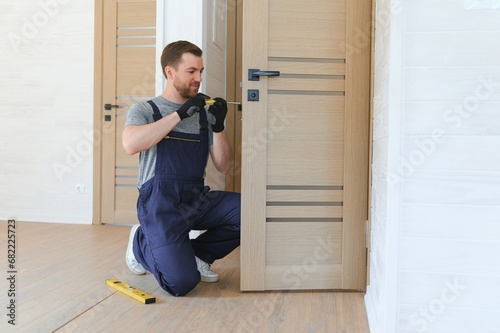 Installation of a lock on the front wooden entrance door. Portrait of young locksmith workman in blue uniform installing door knob. Professional repair service. Maintenance Concept photo