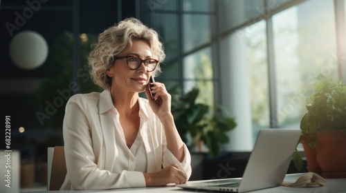 Middle aged European Latin businesswoman having call on smartphone with business partners or clients. Smiling mature Hispanic woman sitting at table talking by mobile cellphone at workplace in office.