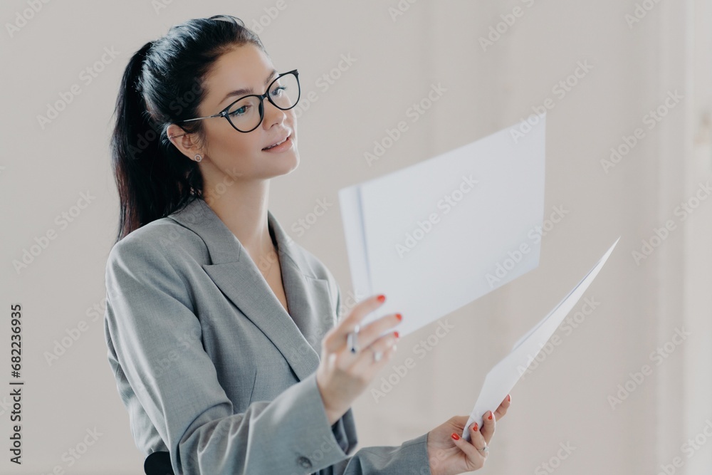 Attentive businesswoman reviewing documents, wearing glasses and a gray suit against a light background. Attentive businesswoman reviewing documents, wearing glasses and a gray suit against a light background.