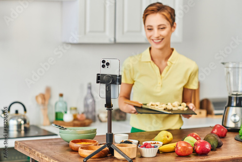 smartphone on tripod near woman with chopping board and fresh fruits with vegetables in kitchen