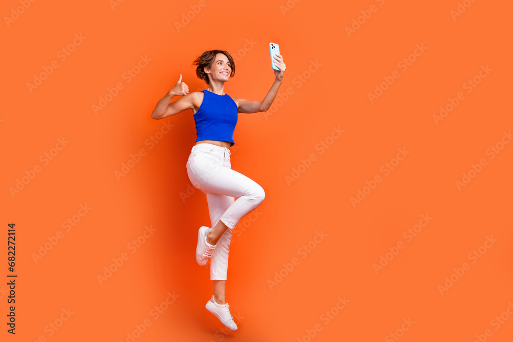 Full size profile photo of attractive sporty girl jump take selfie smart phone show thumb up empty space isolated on orange color background