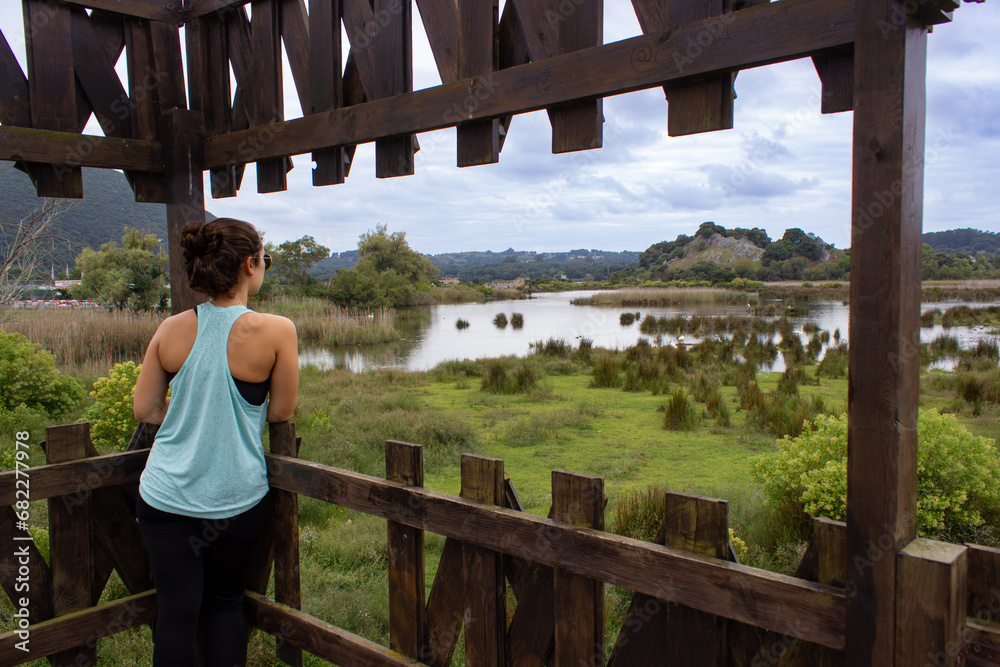 Young woman looking at a beautiful marsh after a run or a walk. long brown hair in a bun. She is wearing sports clothing and a sleeveless t-shirt and standing on a wooden platform. cloudy summer day.
