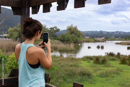 Young woman looking at a beautiful marsh after a run or a walk. Taking a picture with the phone. She is wearing sports clothing and a sleeveless t-shirt and standing on a wooden platform. 