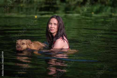 A young beautiful dark-haired girl bathes in a pond with a pit bull terrier dog.