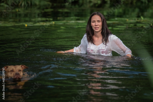 A young beautiful dark-haired girl bathes in a pond with a pit bull terrier dog.