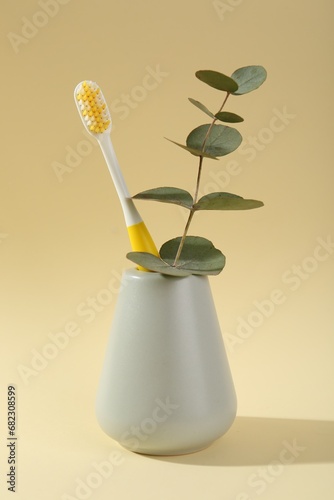 Fototapeta Naklejka Na Ścianę i Meble -  Plastic toothbrush and eucalyptus branch in holder on pale yellow background