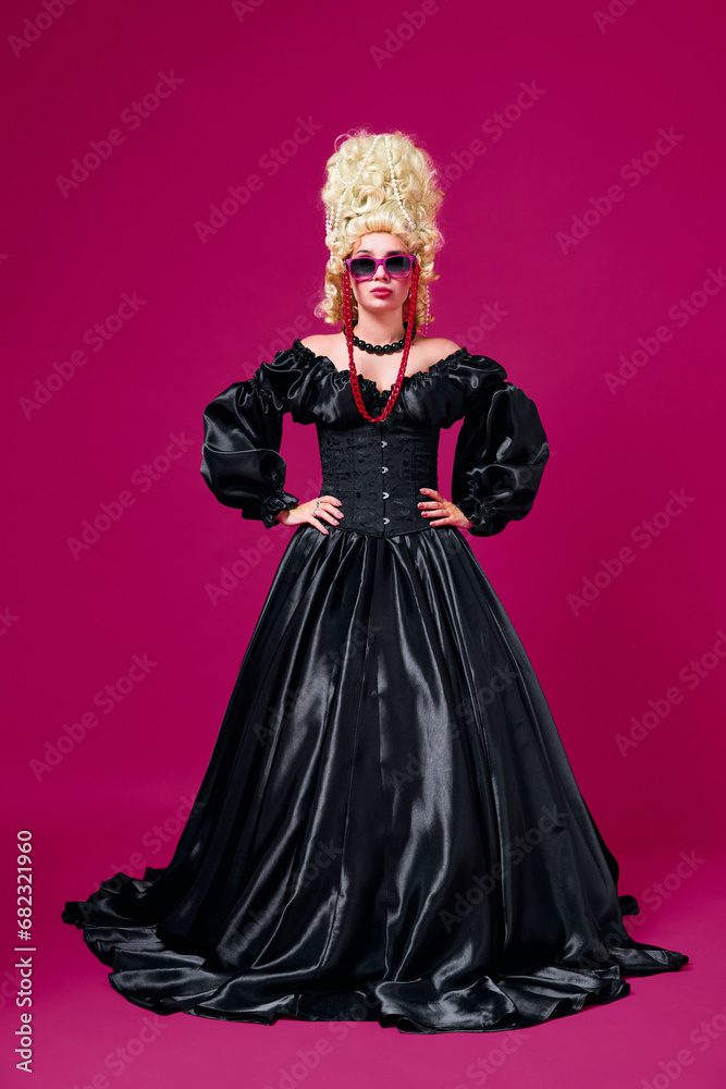 Full length portrait of young girl dressed like medieval person in black old-fashioned dress with modern accessories against magenta background.