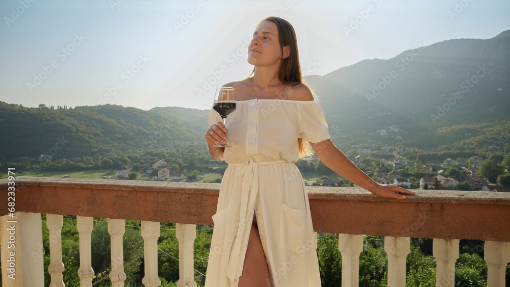 Young woman in a flowing dress relishes a glass of red wine on the balcony or villa terrace, while taking in the magic of a mountain sunset. Young woman in a flowing dress relishes a glass of red wine on the balcony or villa terrace, while taking in the magic of a mountain sunset.