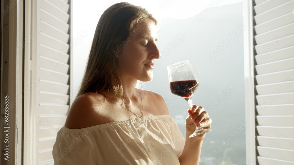 Young woman, standing at an open door on the terrace, gracefully sipping red wine from a glass, as the sun sets over the breathtaking mountains. Tranquility and appreciation of nature Young woman, standing at an open door on the terrace, gracefully sipping red wine from a glass, as the sun sets over the breathtaking mountains. Tranquility and appreciation of nature