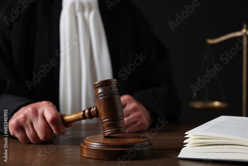 Judge with gavel sitting at wooden table against black background, closeup