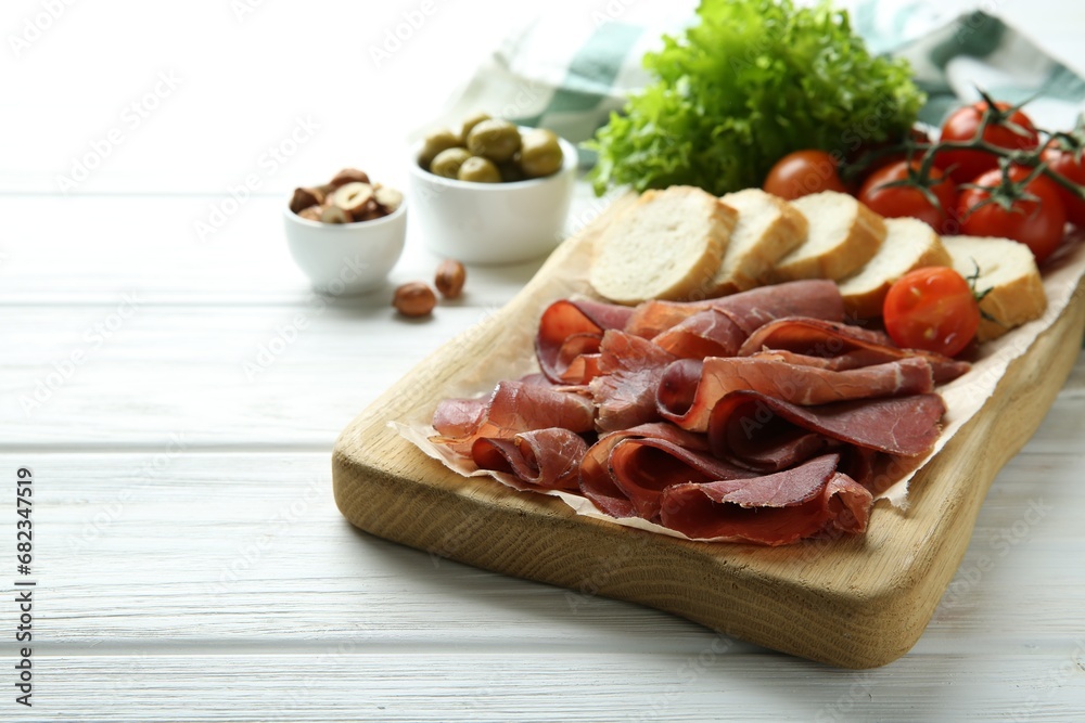 Board with delicious bresaola served with bread and tomato on white wooden table. Space for text