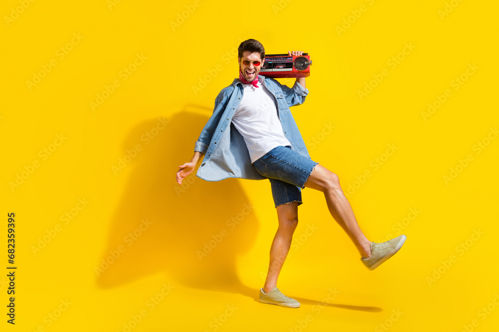 Full size photo of overjoyed guy wear jeans jacket in sunglass hold boombox walk on party empty space isolated on yellow color background