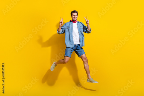 Full size photo of satisfied nice guy wear jeans jacket red scarf jumping showing v-sign isolated on vibrant yellow color background