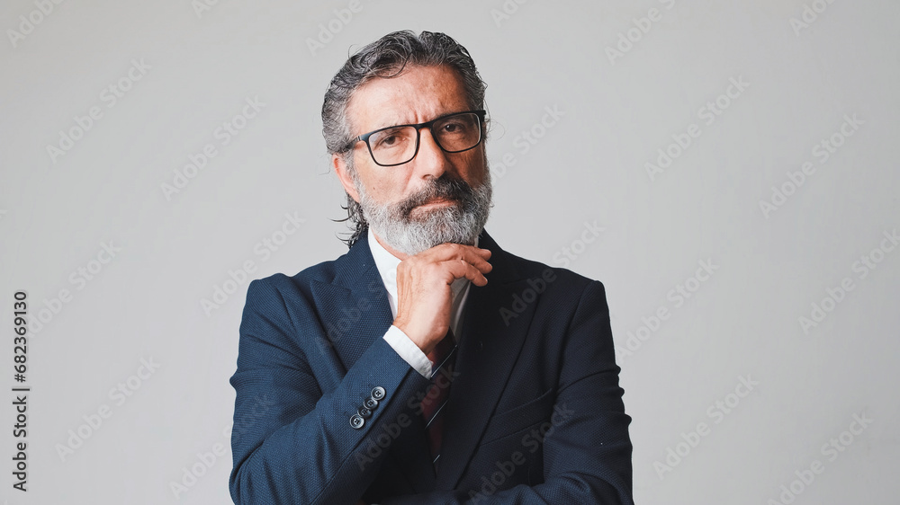 Middle-aged businessman in glasses with gray hair, hand props chin and looking at camera, isolated on gray background in the studio Middle-aged businessman in glasses with gray hair, hand props chin and looking at camera, isolated on gray background in the studio