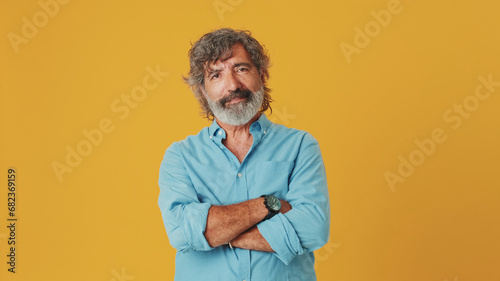 Elderly white-haired bearded man wears a blue shirt, looks at the camera with his arms crossed, isolated on an orange background in the studio