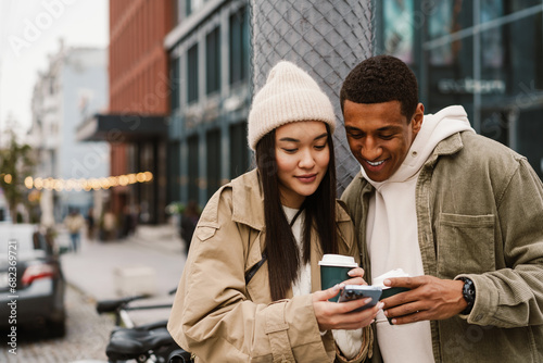 Smiling couple using smartphone and drinking coffee while standing at street photo