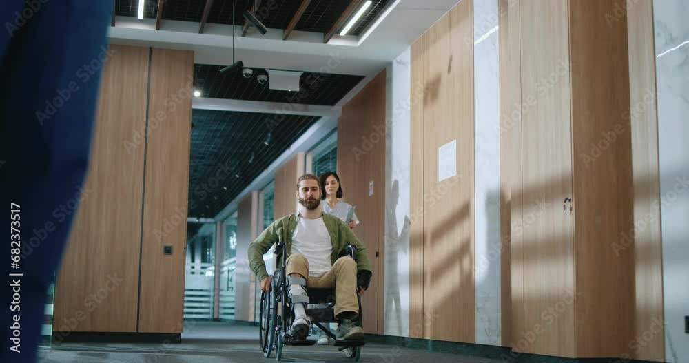 Injured Caucasian handsome patient in wheelchair moving around cozy hospital corridor and greeting medical staff workers. Attractive man with beard recovering in modern clinic. Health care concept. Injured Caucasian handsome patient in wheelchair moving around cozy hospital corridor and greeting medical staff workers. Attractive man with beard recovering in modern clinic. Health care concept.