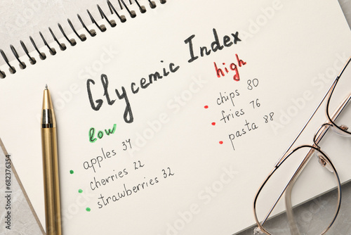 List with products of low and high glycemic index in notebook, pen and glasses on table, top view photo