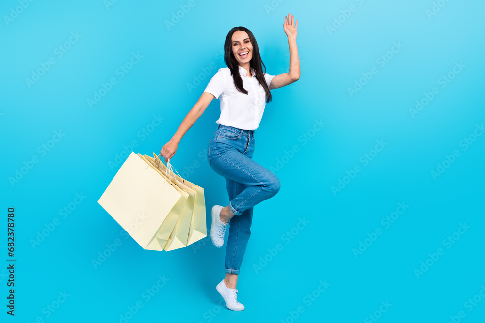 Full length photo of excited funky lady wear white shirt waving arm hi shopping empty space isolated blue color background Full length photo of excited funky lady wear white shirt waving arm hi shopping empty space isolated blue color background