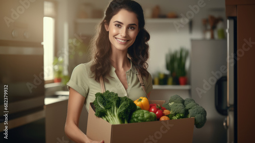 Smiling woman holding a box filled with fresh vegetables in a home kitchen setting. © MP Studio