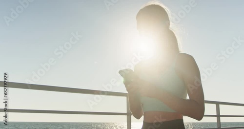 Young female in sportswear walks on sea waterfront after long running and looks at phone to relax. Woman athlete takes short break from run to write message photo