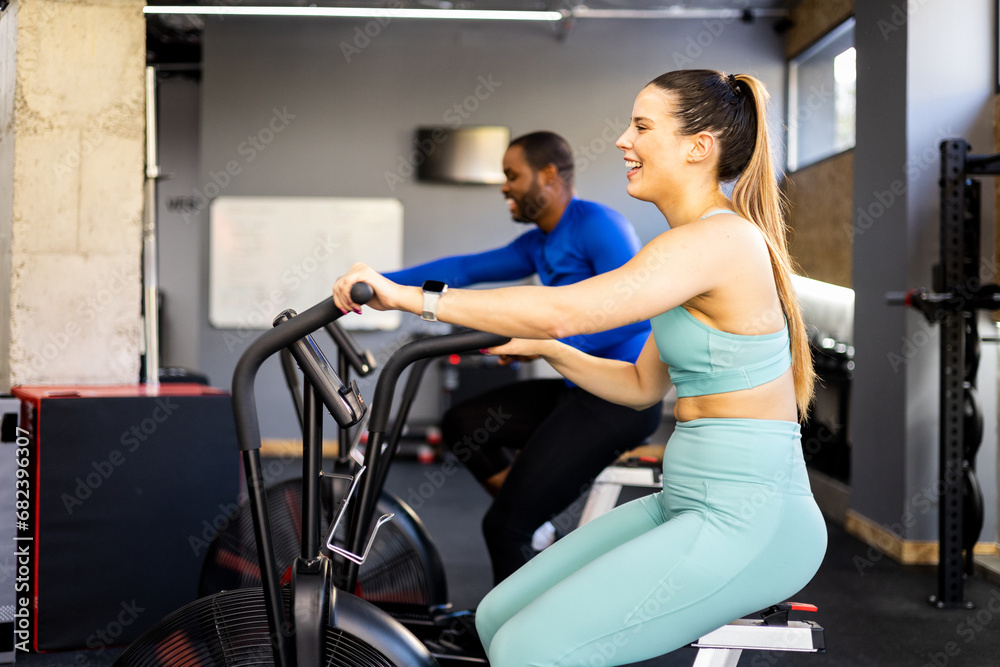 A couple of different ethnicities are doing functional training with stationary air bikes.The young woman is in the foreground with a happy face.Concept of anaerobic classes with air bikes.