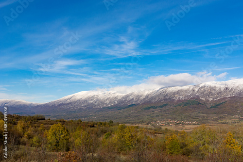 Autumn landscape showcasing trees adorned with golden foliage, with a distant high mountain peak covered in snow. Above, passing clouds in a blue sky on a sunny day in nature