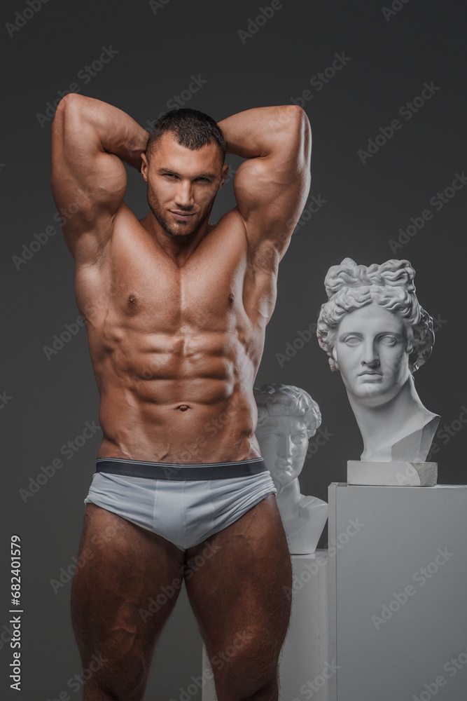Handsome man exuding a rugged charm, with a flawless muscular chest, posing proudly beside ancient Greek statues against a gray backdrop Handsome man exuding a rugged charm, with a flawless muscular chest, posing proudly beside ancient Greek statues against a gray backdrop