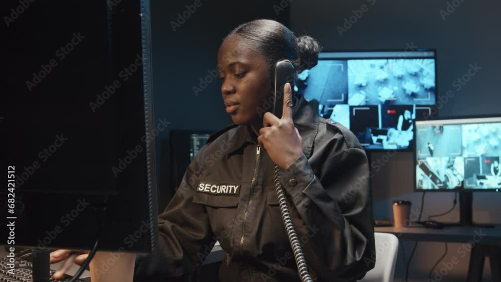 Medium shot of Black female security guard working on computer and answering work call on landline phone in surveillance room with multiple screens of CCTV footage