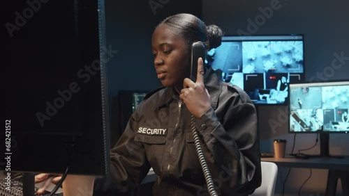 Medium shot of Black female security guard working on computer and answering work call on landline phone in surveillance room with multiple screens of CCTV footage photo