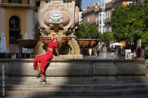 Young, beautiful blonde American woman in a red dress with white polka dots, sunbathing sitting in a wonderful fountain, during her trip to a European city. Concept travel, tourism, destinations.