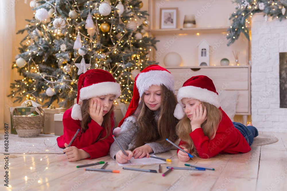 Three sisters write a letter to Santa. The girls are wearing beautiful Santa's helper hats. They have long beautiful hair. They lie on the floor near the Christmas tree Three sisters write a letter to Santa. The girls are wearing beautiful Santa's helper hats. They have long beautiful hair. They lie on the floor near the Christmas tree