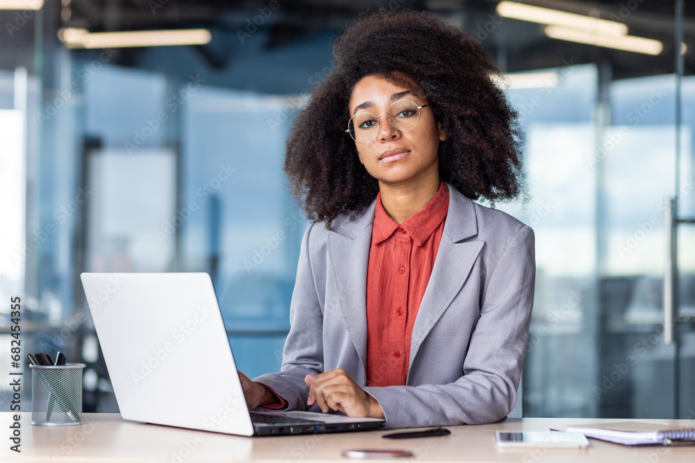 Portrait of a serious and successful African American businesswoman sitting in the office at the desk, working on a laptop and looking confidently at the camera. Portrait of a serious and successful African American businesswoman sitting in the office at the desk, working on a laptop and looking confidently at the camera.