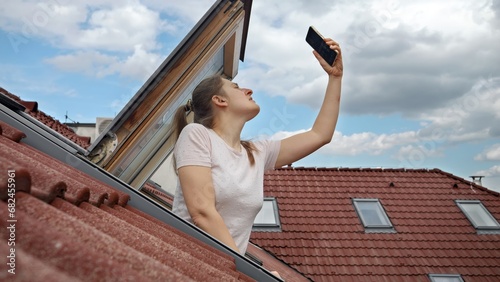 Young woman encounters signal problems with GPS or 5G on her smartphone while looking out of an open attic window, determined to restore connectivity photo