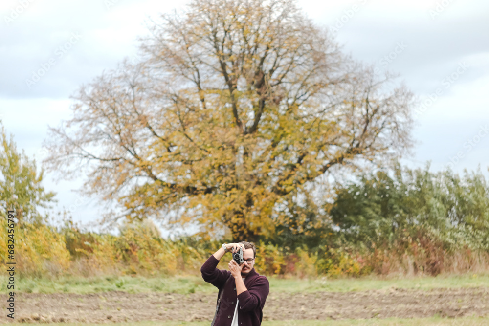 Fashion look, pretty cool young man with retro film camera wearing a elegant sweater outdoors over rural fall background. Design. Photographer or blogger