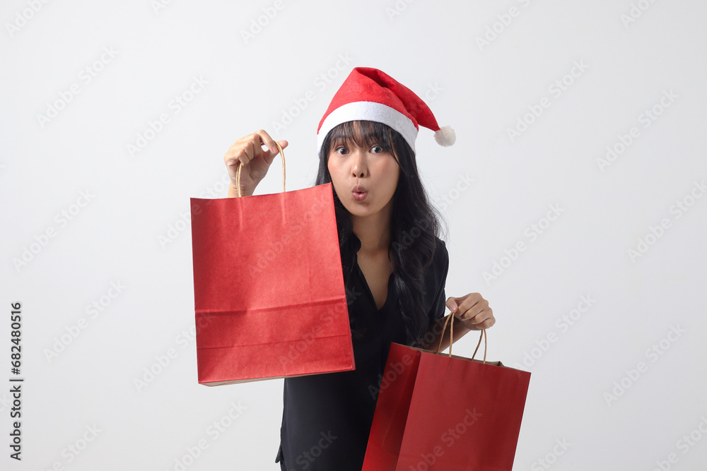 Portrait of surprised Asian woman with red Santa hat feeling happy while purchasing stuff. Excited girl holding shopping bag. New year and christmas concept. Isolated image on white background Portrait of surprised Asian woman with red Santa hat feeling happy while purchasing stuff. Excited girl holding shopping bag. New year and christmas concept. Isolated image on white background