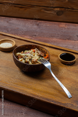 Wok udon noodles with beef and vegetables in a coconut shell plate on a hearth board next to soy sauce and sesame seeds.