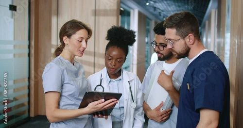 Multicultural group of youthful medical personnel communicating in modern hospital hallway during break. Nurses and doctors sharing experience and spend time using devices online.