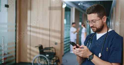 Successful focused professional doctor in uniform using internet to consult online during break in hallway of modern clinic. Productive medical professional using smartphone online. photo