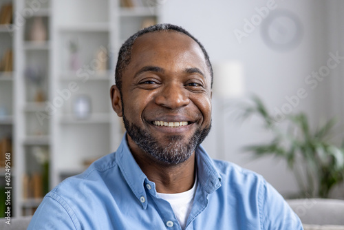 Close-up portrait of mature experienced African American man smiling and looking at camera while sitting on sofa in living room at home.
