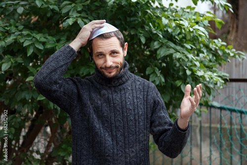 Religious Israeli caucasian man with smile face, holding a kippah, hand to his head. Handsome smiling bearded Jewish holding yarmulke  for wind over his head while looking at camera. Looks surprised