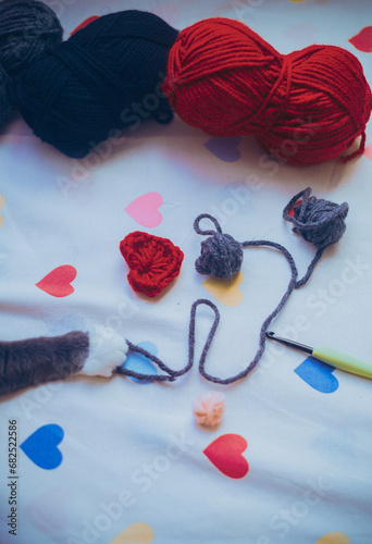 On a cute bed sheet with hearts there are skeins of yarn and a knitting hook, and the cat's playful paw reaches for a ball of wool. A pet interferes with knitting. A cozy hobby.