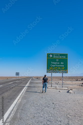 Homem latino de cabelos longos, camiseta preta e jeans fotografando placa de sinalização do Trópico de Capricórnio no deserto do Atacama, Chile.  photo