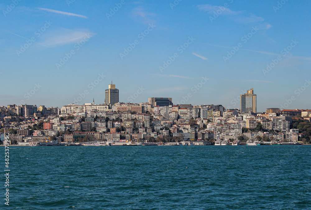 Istanbul's skyline from the Bosphorus: sunny, with a mix of skyscrapers, historic buildings, and several boats on the calm blue water.
