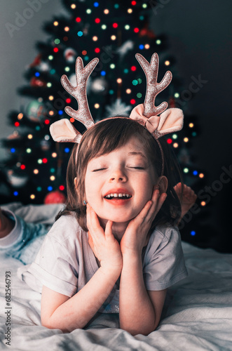 A little beautiful girl is lying on a bed with deer horns on the background of a Christmas tree.