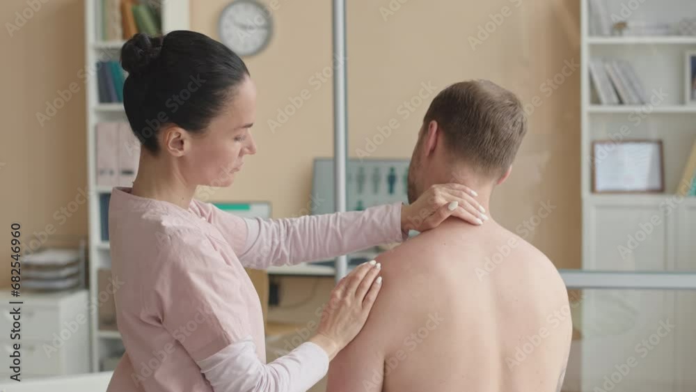 Female physiotherapist palpating arm and shoulder of shirtless male patient while diagnosing muscle problems during checkup in clinic Female physiotherapist palpating arm and shoulder of shirtless male patient while diagnosing muscle problems during checkup in clinic