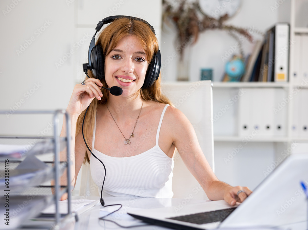 Naklejka premium Portrait of positive young woman wearing headset, using laptop. Call center worker sitting at desk in office.