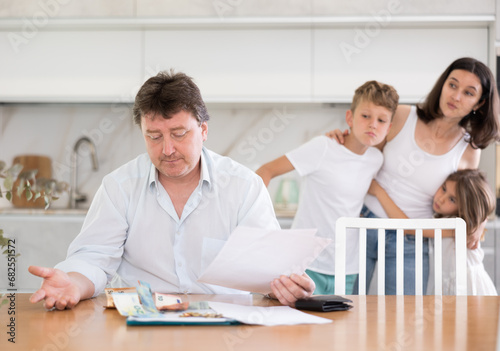 Sad father looks at bills and counts money. Upset wife with children in the background